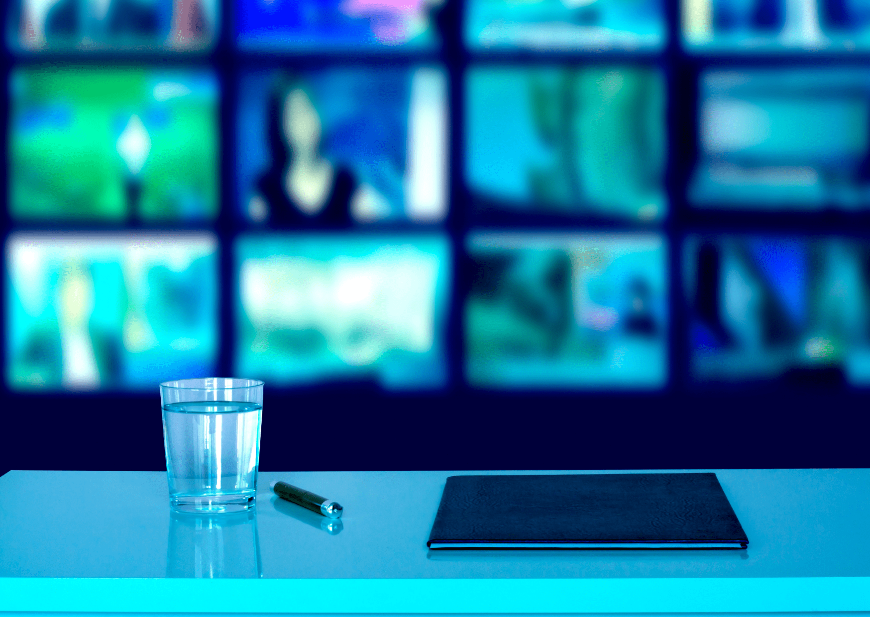 A notebook, pen and glass of water on a table, with a background of television screens.
