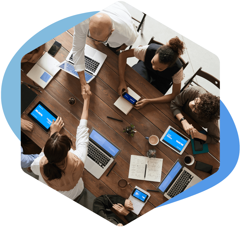 team sat around a table, on laptops, shaking hands. having a team meeting.
